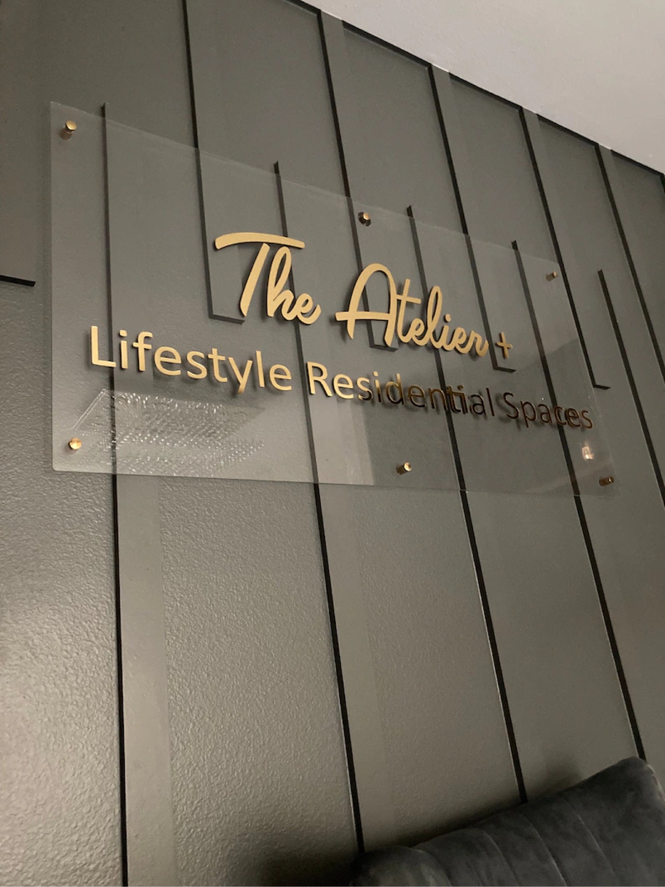 Gold letters reading The Atelier+ Lifestyle Residential Spaces on a clear acrylic panel